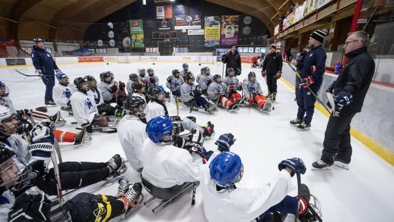 About four officials speaking in front of a group of Para ice hockey players