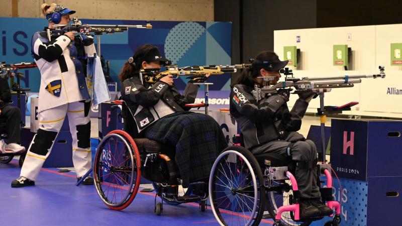 A standing woman and two women in wheelchairs competing in a rifle shooting range