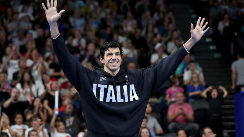 A male Para swimmer wearing a hoodie that says Italia raises his arms in front of a crowd