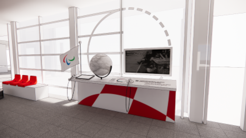 A wheelchair accesible desk with a tactile globe, a screen and handsets are placed next to the IPC flag and a bench.