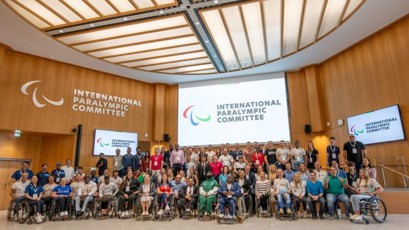 A group photo taken during the IPC Athletes' Forum - more than 70 athletes and officials are smiling in front of a screen with the IPC logo