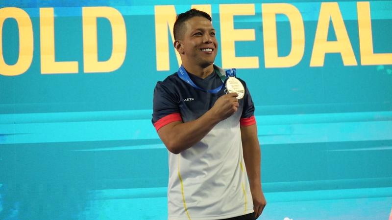 A man showing his medal with a backdrop with the words gold medal behind him