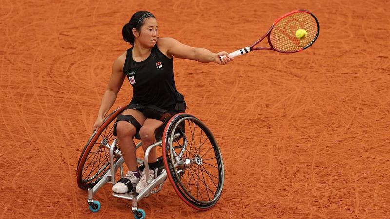 A female wheelchair tennis player plays a forehand on red clay court