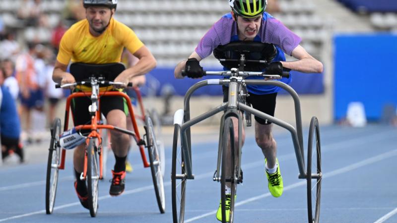 Two athletes are racing on a blue athletics track. They are running using a metal frame with three wheels