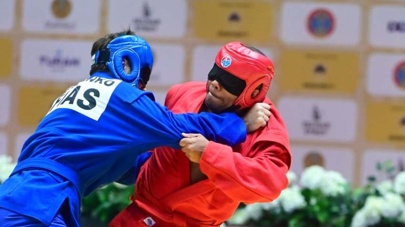 Two male athletes wearing blindfolds are wrestling - one athlete is wearing a red uniform and helmet while his opponent is wearing a blue helmet and gear