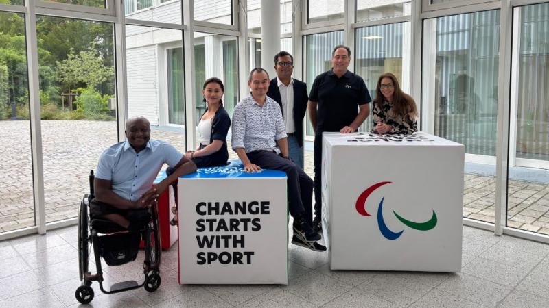 Six people pose for a photograph in front of two blocks that say &quot;Change Starts with Sport&quot; at the IPC Campus in Bonn, Germany
