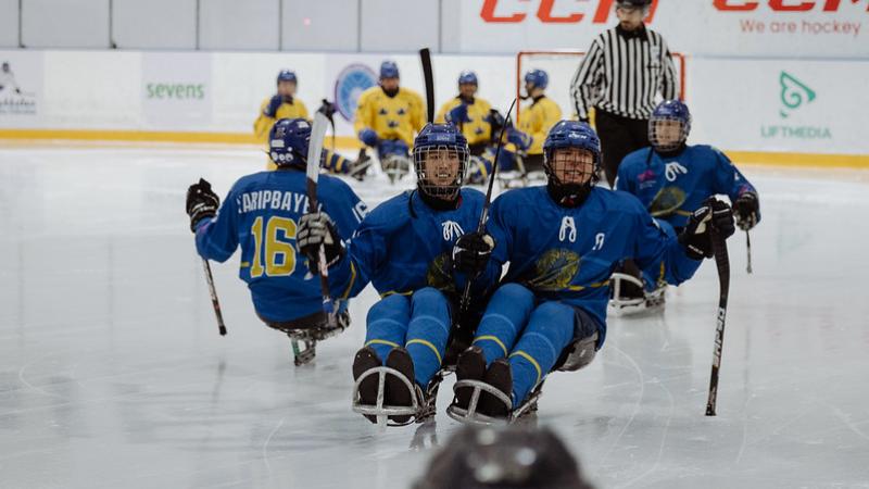 A group of four Kazak Para ice hockey players on ice with an official and four other players in the background