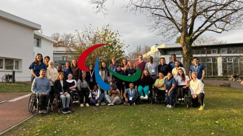Twenty retired Para athletes take a photo in front of the Agitos symbol at the IPC Campus