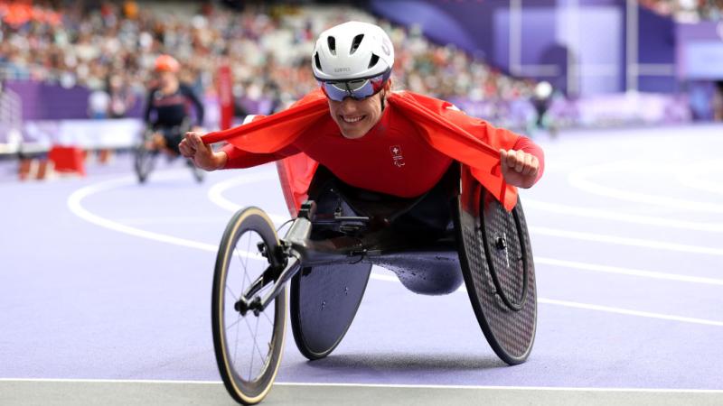 A female wheelchair racer dressed in a red gear and white helmet is holding the Swiss flag after a race