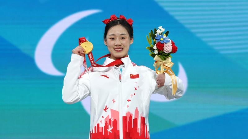 A female athlete with red ribbons on her head smiles on the podium while holding a flower bouquet and a gold medal