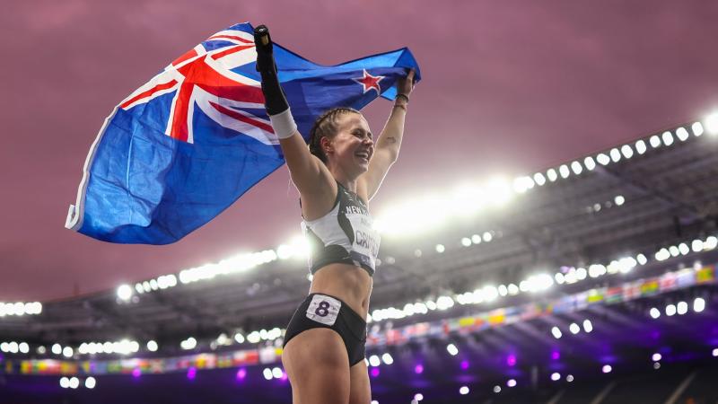 A female Para athlete wearing a track suit is holding New Zealand's flag in an outdoor stadium