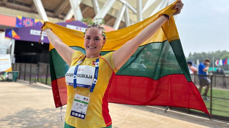A female athlete wearing a green, yellow and red athletic gear is holding the Lithuanian flag in front of a stadium