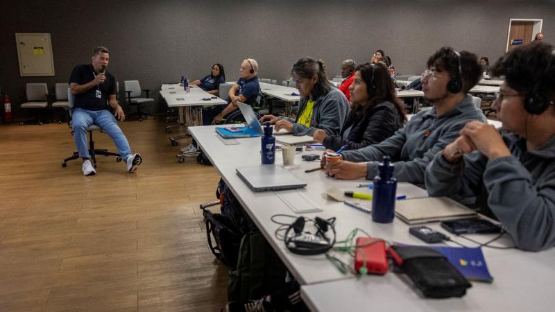 About 10 people sit in a classroom, listening to a male official speak 