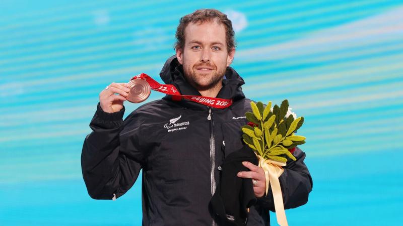 A male Para alpine skier celebrating his win, by holding a bronze medal and flowers