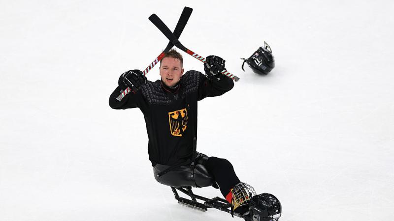A male para ice hockey player in black crosses his sticks above his head