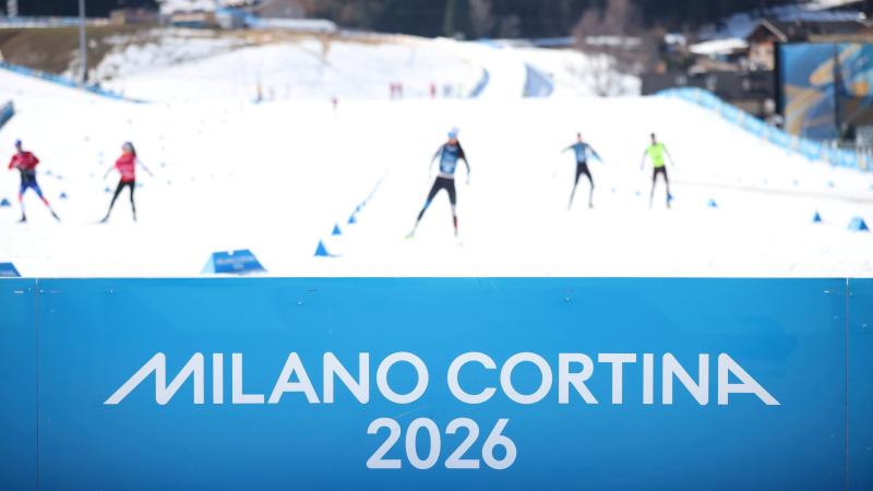 A banner with Milano Cortina 2026 written on it, while in the back para biathlon athletes are training