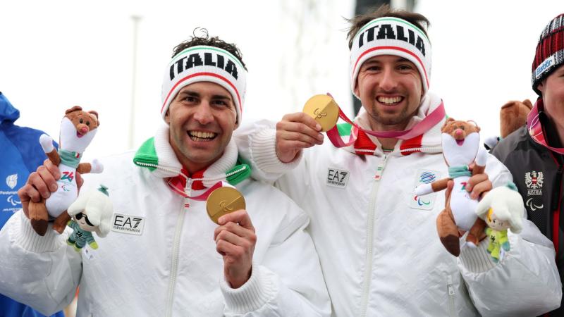 One male Para athlete and his male guide celebrating while holding their gold medals in one hand and the Milo Paralympic mascot in the other hand, wearing headbands with the words ''Italia'' and smiling at the camera