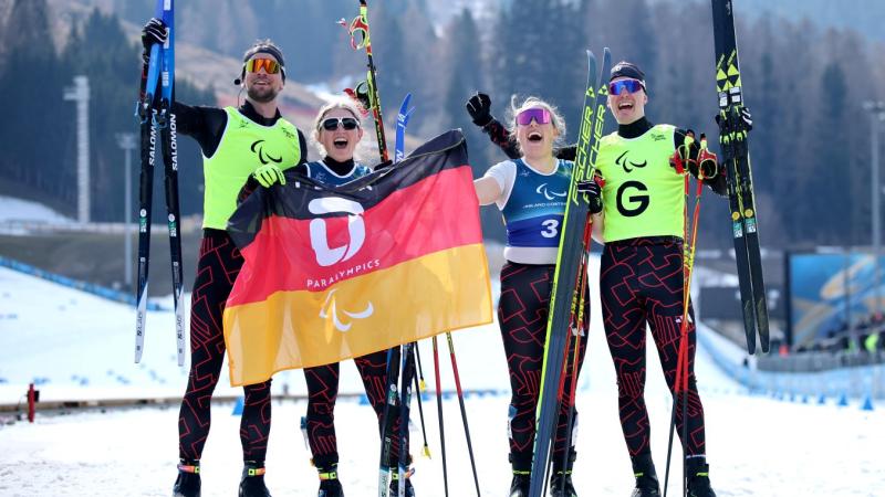 Two female Para cross-country skiers celebrating after competition waving their skis and the German flag, alongside their male guides