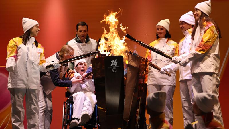 Seven torchbearers wearing a white uniform are lighting a cauldron 