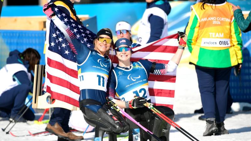 Two female sit-skiers pose for a photo by holding the flag of the USA