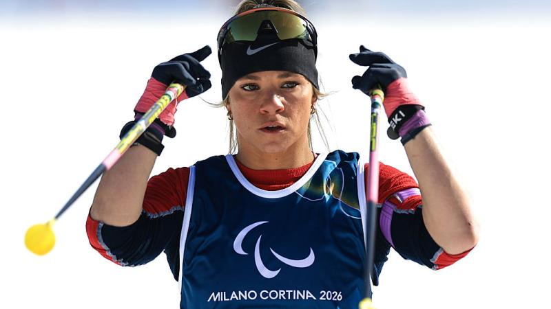 A female athlete wearing a blue bib is holding ski poles