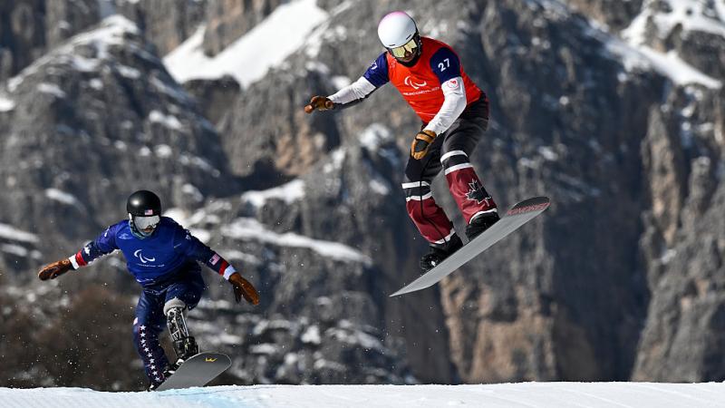 A male snowboarder in red leads a male snowboarder in blue against a mountain backdrop