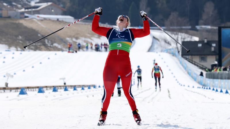 A female Para cross country skier celebrating pointing at the sky, as she crosses the finish line