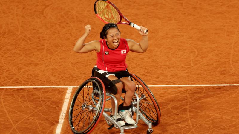 A female wheelchair tennis athlete is celebrating on a red clay court