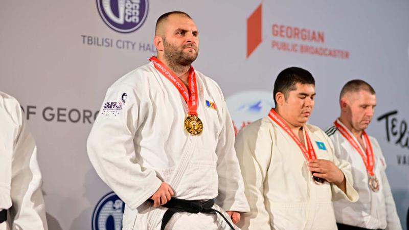 a male Para judoka with a Moldovan flag on his chest stands on the podium with a gold medal around his neck 