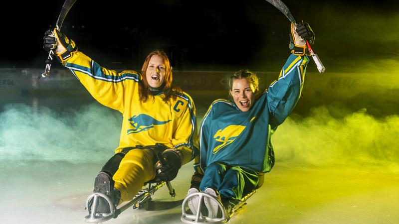 Two female Para ice hockey players with Team Australia jerseys raising their sticks on ice