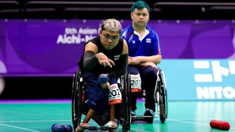 A male athlete, who is in a wheelchair, is throwing a blue boccia ball on the court