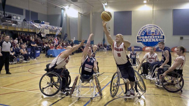 A picture of a man in a wheelchair shooting a Basketball ball between 2 other men in a wheelchair.
