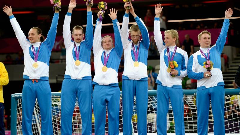 Six men in training suits standing in a row, waving and celebrating