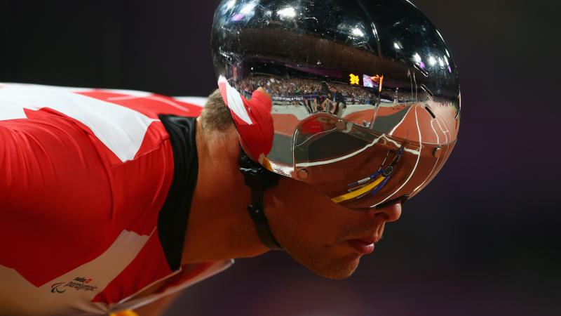 A man with a silver helmet and red jersey leans forward on his racing wheelchair