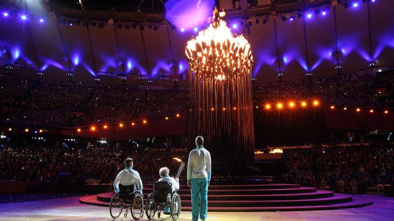 A picture of one man and woman in wheelchairs and one man standing watching a lightened cauldron during a ceremony
