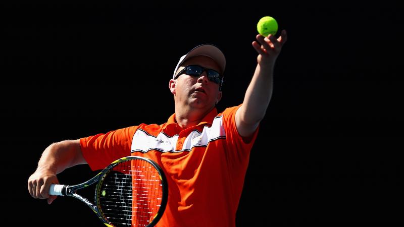 A picture of a man in a wheelchair serving during a tennis match