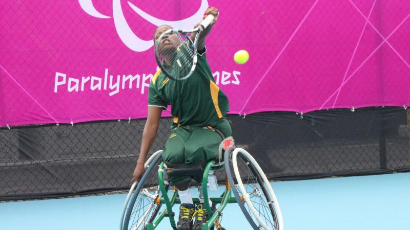 A picture of a black woman in a wheelchair serving during a wheelchair tennis match.