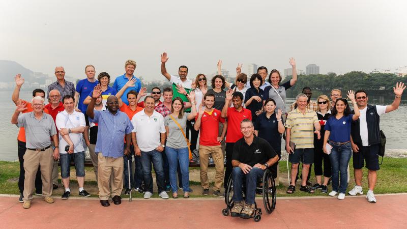 A group of people pose for a photo, waving in front of a body of water.