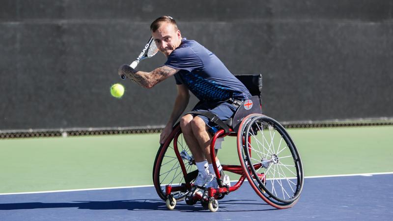A man in a wheelchair swings back a tennis racket in preparation of hitting a backhand.