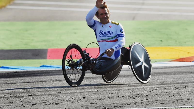 A man in a handcycle waves to spectators from a dirt track.