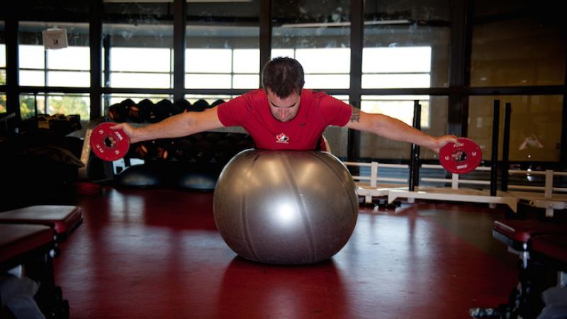 An athlete lies on his stomach on a stability ball with both his arms stretched out to the sides holding weights.
