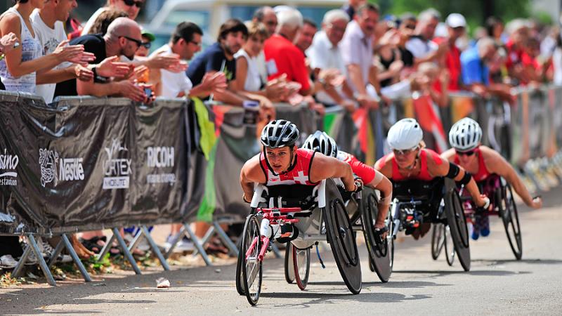 A woman looks up as she races in a wheelchair at the front of the pack with fans along the side of the road.