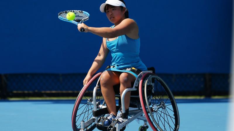 A woman in a wheelchair hits a tennis ball as a backhand.