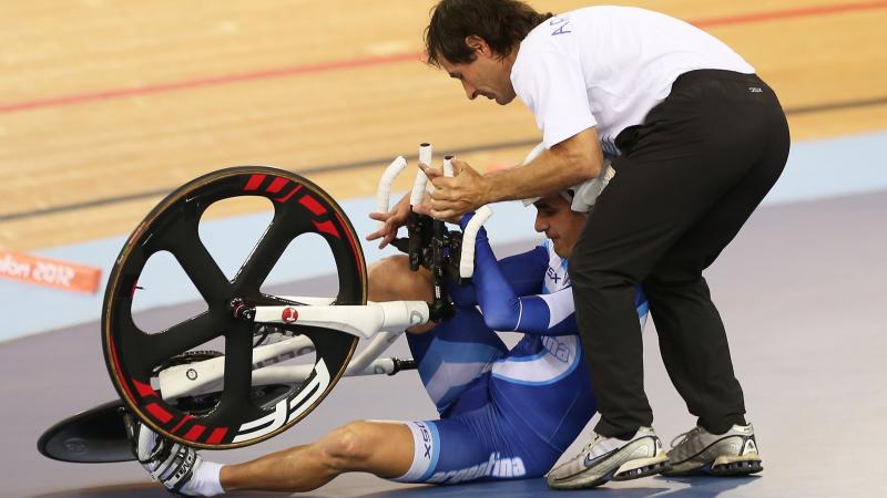 A man sits beside the track trying to get up with his bike with another man assisting him.