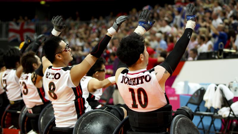 Wheelchair rugby players shown from behind raise their arms and wave to a crowd.