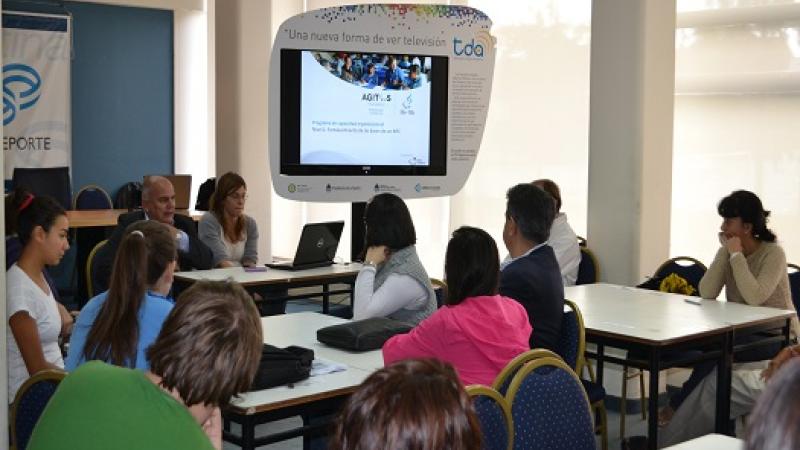 People sitting on chairs around tables, looking at a screen. Classroom atmosphere