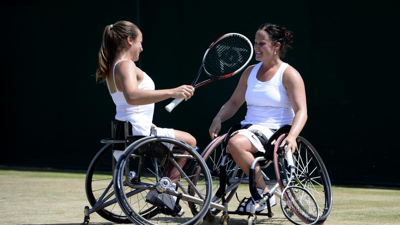 Two women in wheelchairs look at each other smiling on a grass tennis court.