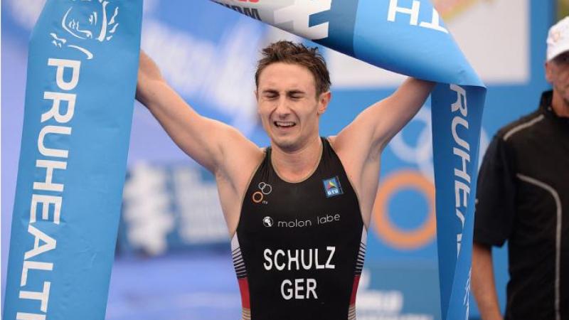 A para-triathlete closes his eyes as he crosses the finish line and raises the finish line banner up in the air.