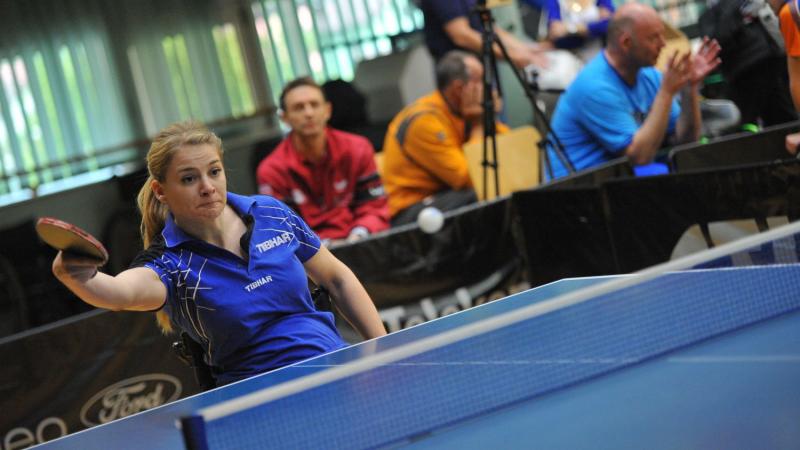 Women in wheelchair in behind a table tennis table batting the ball.