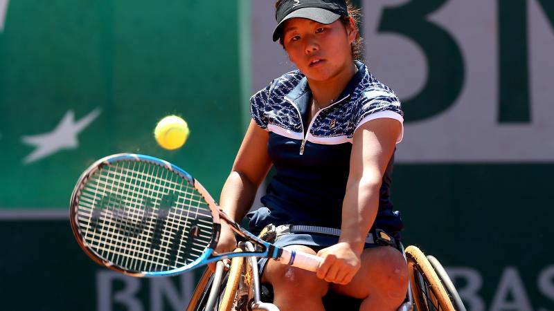 Woman in wheelchair on a tennis court returning a shot.